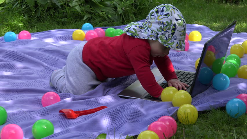 Infant baby left alone type play with laptop computer between colorful balls on plaid in park. Static shot.
