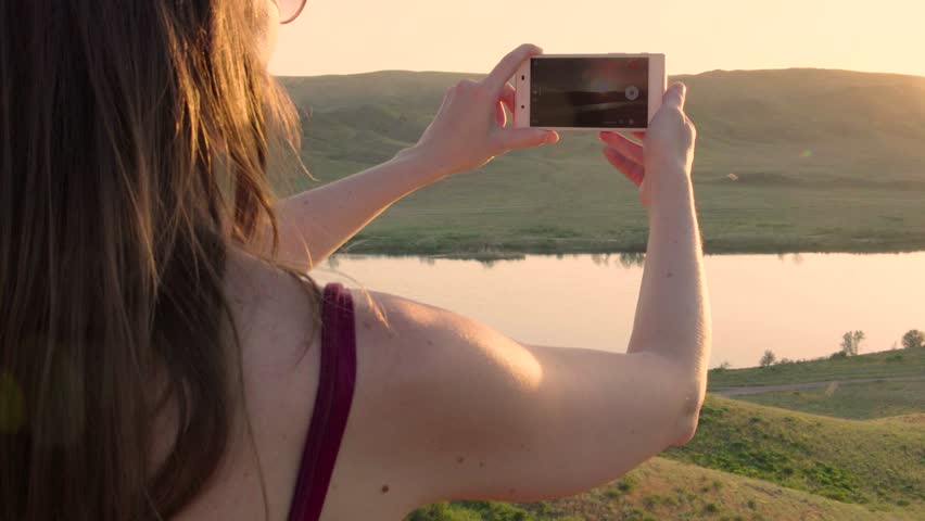 Close up from the back of girl or woman taking photo of sunset lake view landscape river at the background with her smart phone. Almaty.