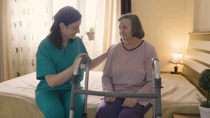 Friendly caregiver helping senior woman getting up from bed and walk with a walker. Home or hospice nursing and assistance concept. 4K footage at 60fps. | Shutterstock HD Video #27546529