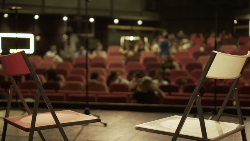 Red and white chairs on stage. Point of view from stage before the start of the show. Red theater seats, people and light in cinema or concert hall. audience enters before the show