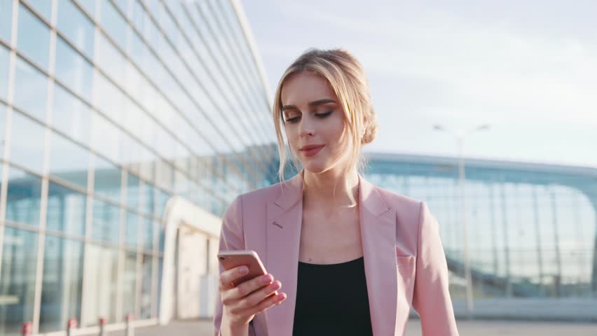 Self-confident hot young blonde girl in an elegant pink jacket walks by the airport terminal and uses her cellphone, smiles happily to the text messages, looks around. Stylish outfit, modern woman.