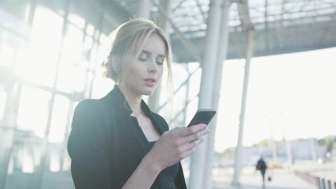 A beautiful blonde woman in a formal black outfit stands by the airport entrance, uses her cell phone, gets the text message, being surprised, smiles happily and looks around.