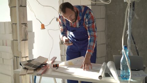 Male worker working with smartphone and blueprints during renovation at new home
