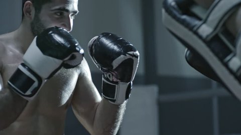 Medium shot of shirtless MMA fighter in boxing gloves hitting and leg kicking punching pads held by his sparring partner