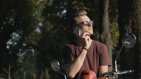 Young attractive man wears sunglasses on retro motorbike speaking phone in park, hipster concept