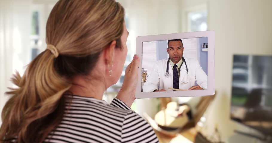 Caucasian woman video calling black male doctor on tablet inside bedroom. Female or patient shown from behind speaking with medical professional through video chat from home. 4k Royalty-Free Stock Footage #27599476