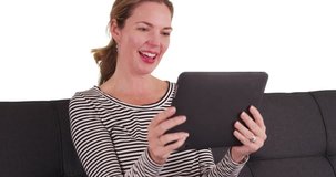 Caucasian woman using tablet to make video call, waving at screen on white background with copyspace. Smiling female sitting on couch or sofa, having video chat in studio with copy space. 4k