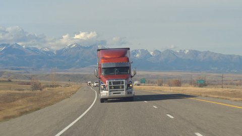 CLOSE UP: Freight container semi truck hauling goods, cars, SUVs and pickups traveling along the busy traffic highway running through farmland countryside with snowy mountain-peaks in the background