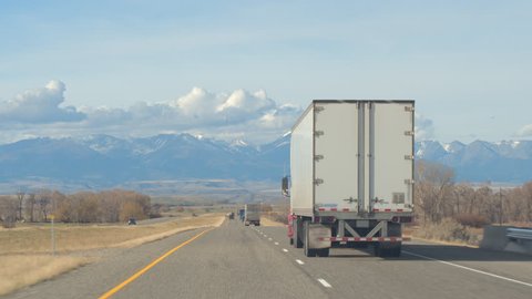 FPV Freight semi trucks transporting goods, personal cars on a road trip, people in SUVs traveling along the busy traffic highway running through farmland countryside towards the Rocky Mountains Range