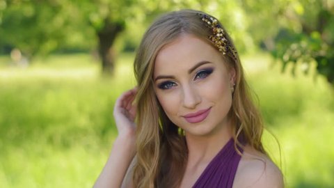 Portrait of a beautiful girl with bright make-up. Caucasian blonde model posing and looking at camera in lilac or purple dress in green garden near apple fruit tree. Slow motion shot.