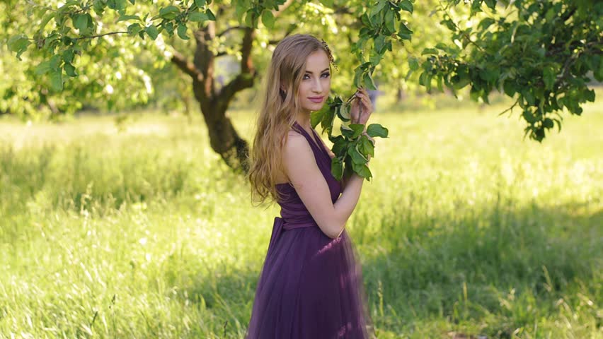 Portrait of a beautiful girl with bright make-up. Caucasian blonde woman with a bouquet of flowers posing and looking at camera in lilac or purple dress in the garden. Slow motion.