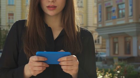 Cute brown haired girl messaging to somebody on her phone outdoors. Close up of female hands typing on touchscreen of smartphone. Pretty young woman smiling while texting on her cellphone