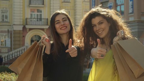 Two female friends approving their shopping in the city. Pretty caucasian girls holding bags on their shoulders. Attractive young women raising their thumbs up