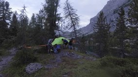 Aerial drone flight over four people setting hanging tent camp near lake. Group of friends summer adventure journey in mountain nature outdoors. Travel exploring Alps, Dolomites, Italy. 4k video side