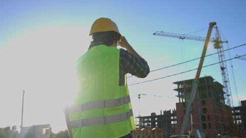 Back view of architect with black curly hair in shirt and green vest looking on construction process, putting on head the helmet on unfinished construction background.