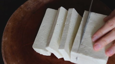 Close up of female hands cutting tofu (made of soy beans) in big slices on round wooden board. Tofu is mainly used in Asian and vegetarian cooking. Table top view.