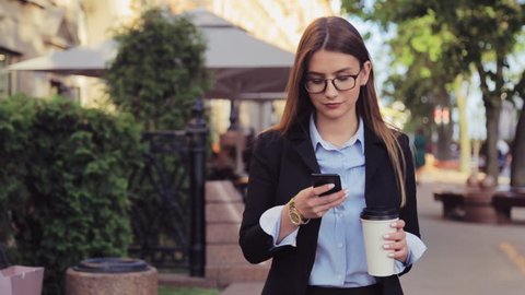 Young business woman is walking on the street and drinks coffee and using smartphone at lunch break.