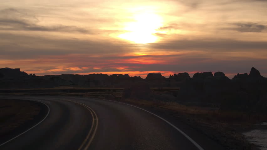 Amazing sunset sky behind the rocky sandstone formations in Badlands National Park. Winding road through Badlands grassland desert in South Dakota at reddish sunrise. Road trip across United States
