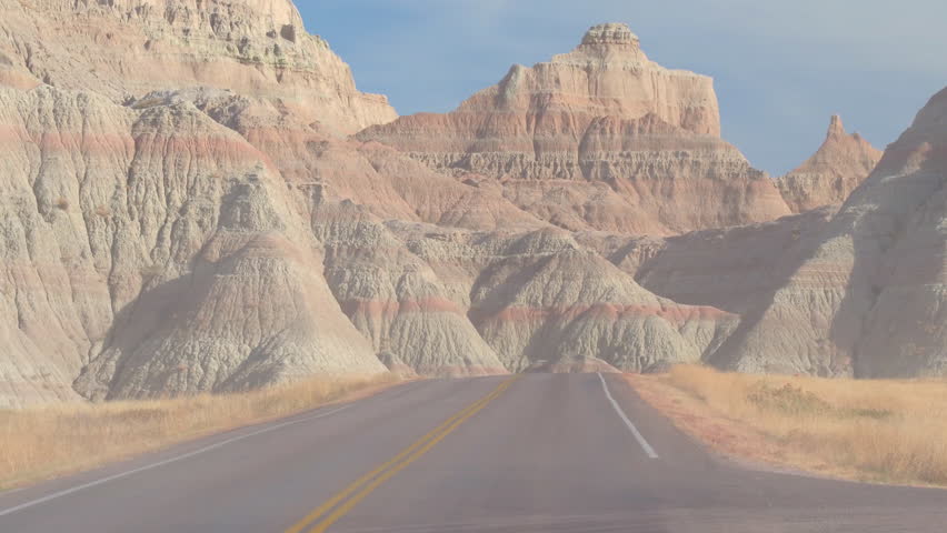FPV: Driving along the dusty road towards picturesque Badlands landscape with rocky sandstone formations. Traveling across the Badlands grassland desert in South Dakota. Road trip across United States