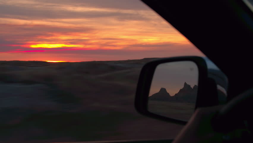 Road trip through Badlands National Park in the Great Plains at amazing reddish sunset. Driving through vast grasslands at beautiful sunrise, sandstone mountains reflecting in wing mirror of a car