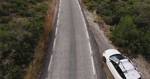 Aerial footage from drone following young unexperienced driver learning how to drive small white hybrid car on mountain road with switchbacks and agricultural forest scenery, obeys road signs