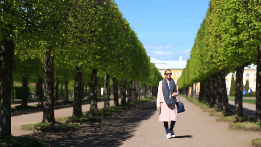 A girl in a pink coat with sneakers and sunglasses walks along the park alley, with a phone in her hand