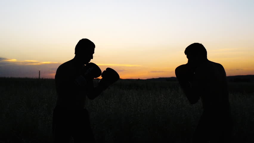Boxing training outdoors. Opponents in boxing gloves silhouette sparring in field at sunset. Fighter kicking with a turn.