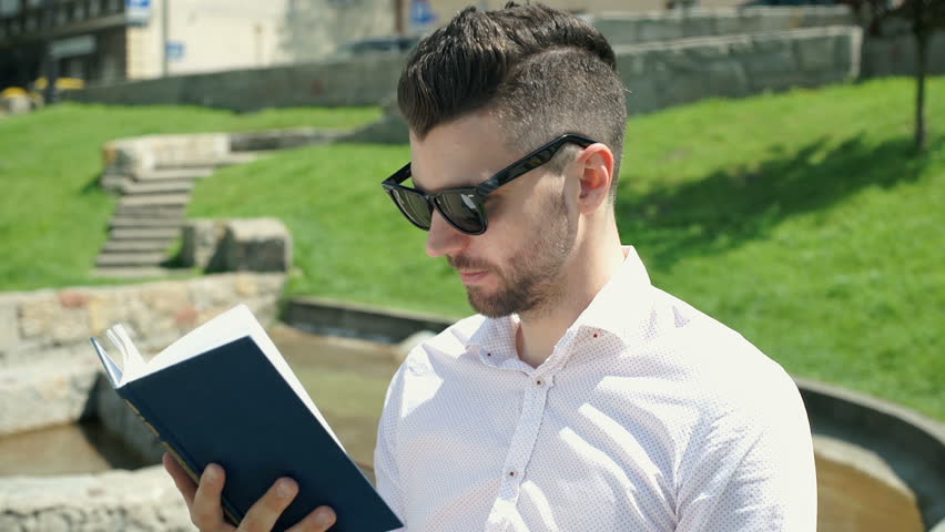 Handsome man wearing sunglasses and reading book in the park, steadycam shot
