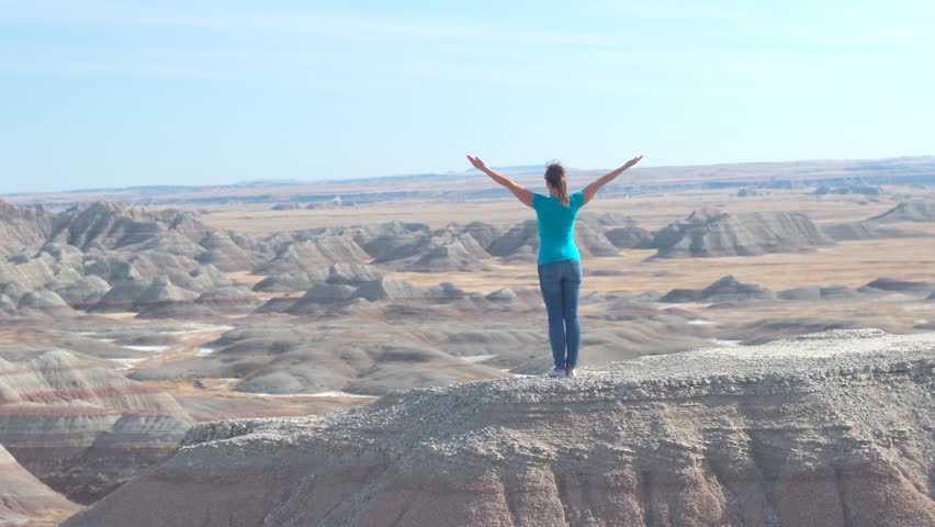 SLOW MOTION Happy woman hiker standing on top of sandstone mountain in Badlands National Park with her arms raised up towards sunny sky enjoying and relaxing. Traveler exploring Badlands wilderness