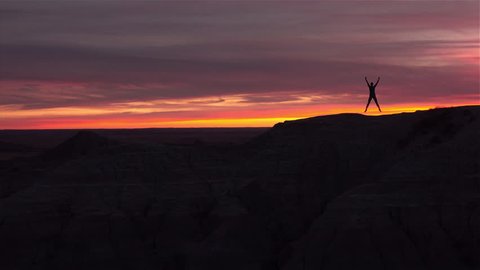 SLOW MOTION: Cheerful woman jumping up with arms raised at pink sunset. Silhouette of female celebrating reaching high rocky mountaintop against pink reddish sunrise sky. Happy girl jumping in success