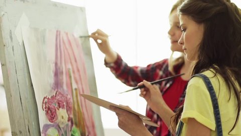 art school, creativity and people concept - student girls or friends with easel, paintbrushes and palettes painting still life at studio