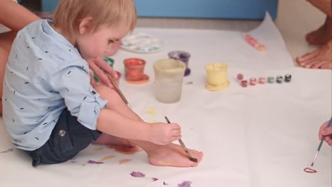 Cute little baby boy painting his feet on a large blank white paper