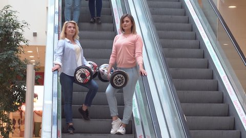 Two attractive girls descend on an escalator in the mall and are holding a hyroscooter in hand