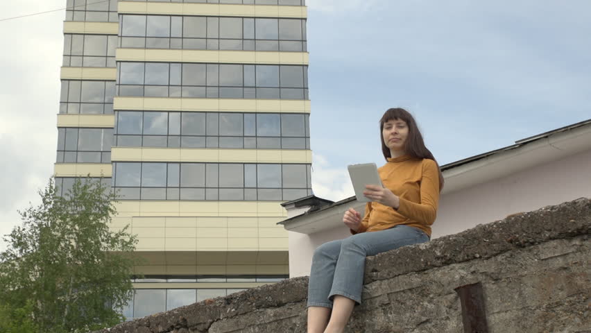 Young happy woman brunette takes self-portrait on a tablet sitting on the edge of the roof on a background of a tall building on a summer day.