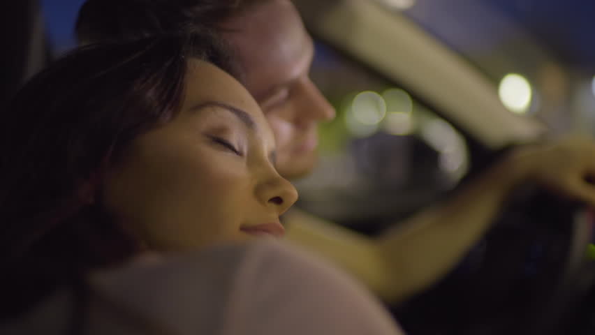 Couple Drive Around City At Night, Woman Leans Against Her Boyfriend's Shoulder, Closes Her Eyes, Sleepy, Then Opens Them And Smiles (Slow Motion)
