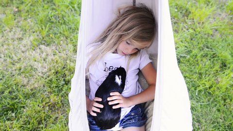 Child blond girl sitting or lying in a hammock and holding her guinea pig pet animal.