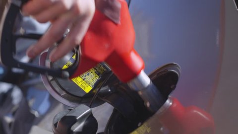 Closeup slow motion shot of male hand taking off fuel cap and inserting nozzle into car tank at gas station; woman drinking coffee in background