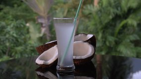 Closeup of coconuts and coconut water with straw on the black glass table isolated over blurred palm trees background - video in slow motion