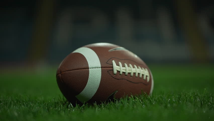 Brown american football ball lying on green grass on stadium. Then player picking it in slowmotion. Close up