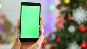 Closeup of female hands holding black modern smartphone in hands at glowing Christmas home interior. Adult woman uses digital device with green screen touching it with finger. Real time full hd video.