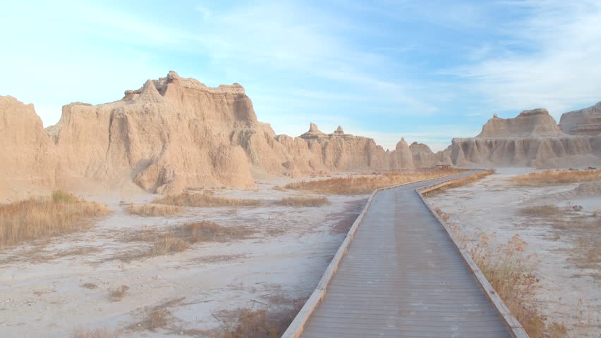 FPV: Walking along the empty boardwalk leading through stunning Badlands desert landscape on sunny morning. Wooden trail going through rocky mountains and grassland of Badlands National Park