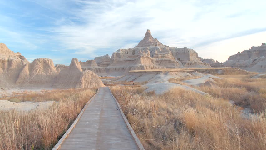 FPV: Walking along the empty boardwalk leading through stunning Badlands desert landscape on sunny morning. Wooden trail going through rocky mountains and grasslands of Badlands National Park