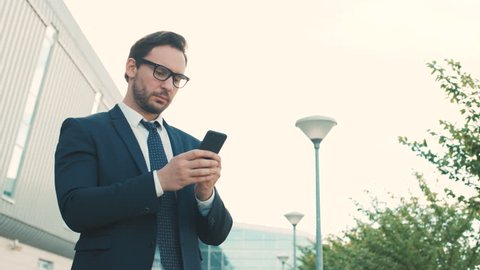 Attractive young businessman using smartphone while standing outdoors near office center building. Man reading, texting on mobile phone. Close up