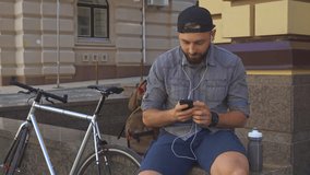 Handsome bearded cyclist having a video chat on his smartphone on the street of the city. Young caucasian man sitting on the edge near his bicycle. Attractive brunette guy in cap waving his hand for