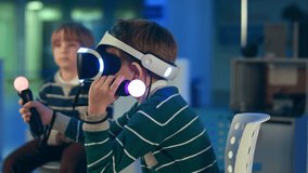 Little boy in vr headset playing virtual reality game with controllers while another boy waiting for his turn