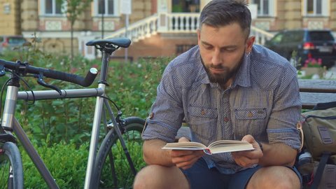 Attractive bearded cyclist reading book on the bench. Young caucasian man looking at the page of some literature. Handsome brunette guy sitting near his bike against background of public building
