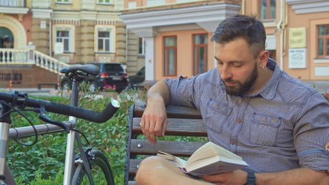 Young brunette cyclist turning the page of the book on the bench. Handsome bearded man holding some literary work in his hands. Attractive caucasian man relaxing by reading near his bike