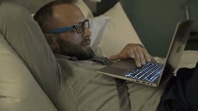 Young businessman watching movie on laptop on bed in hotel room at night
