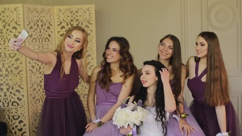 Bridesmaids taking a selfie on a smartphone and laugh during the morning of the bride in a beautiful photo studio. Beautiful girls in purple wedding dresses have fun in the studio.