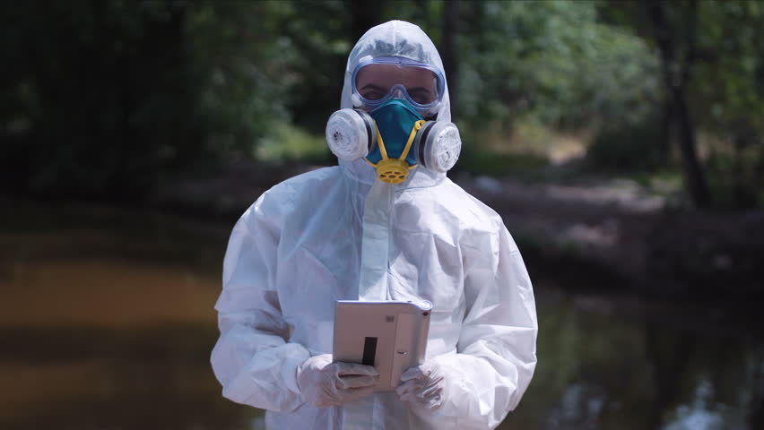 Anonymous female biologist in white protective suit posing with tablet in hands on background of nature.